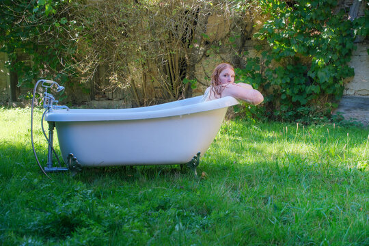 Redhead woman bathes in vintage tub placed outdoors on grass surrounded by greenery while looking back creating unusual scene combining nature and hygiene with quiet reflective mood
