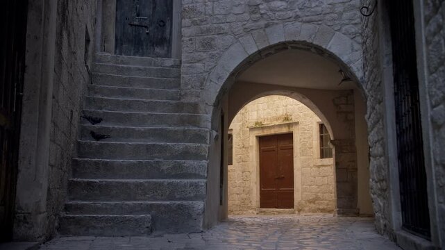Quiet morning street scene in Trogir Croatia with sun partially illuminating building facade with wooden door, while foreground is still in darkness, with two pigeons on the staircase 