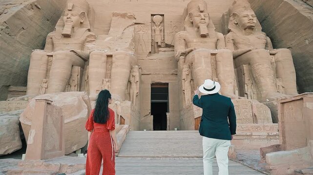 american young man traveler wearing white hat with his wife with red dress exploring ancient Egyptian abu simble temple with background of ramsses ii statues. Egypt.