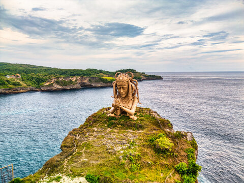 Aerial view of a giant female statue on a grassy cliff overlooking the turquoise ocean and rugged coastline in Nusa Penida, Bali, Indonesia.