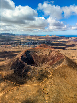 Aerial view of the volcanic crater of Calderon Hondo with its reddish slopes and surrounding arid plains under a cloudy sky in La Oliva, Canary Islands, Spain.