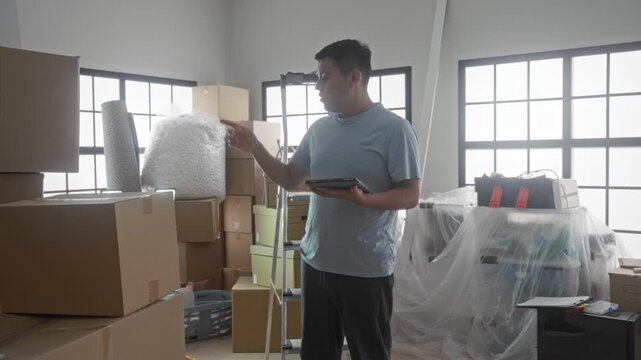 Man holding tablet points to stacked moving boxes and bubble wrap in a new home building while standing by a ladder; determination planning.