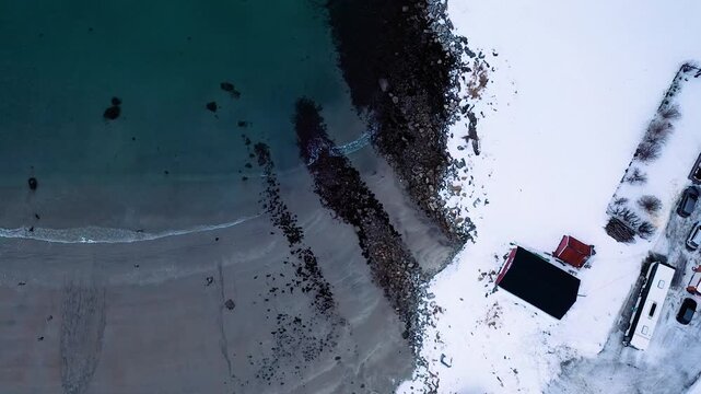 Top Down Drohnenaufnahme von arktischer K&uuml;ste mit Fischerhaus Wellenbrechern und t&uuml;rkisfarbenem Meer in Norwegen