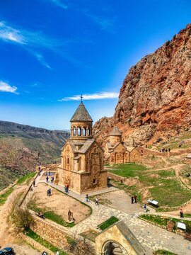 Aerial view of Noravank monastery complex nestled against towering red cliffs with tourists walking on stone paths under a clear blue sky in Areni, Vayots Dzor Province, Armenia.