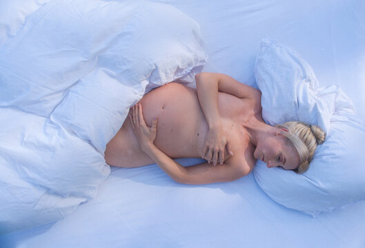 Pregnant woman sleeps on white bed gently holding belly with soft blue tones and light bedding creating intimate peaceful scene focused on rest and maternal connection