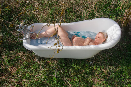 Pregnant woman relaxes in vintage bathtub outdoors surrounded by grass and branches holding belly while resting in water creating unusual natural setting with intimate serene mood