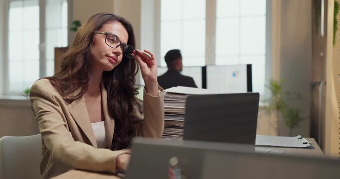 Woman with glasses at laptop in office. A business analyst pauses to focus and ease eye strain from computer work under office light, at a tidy desk. Concise concept of digital eye strain.