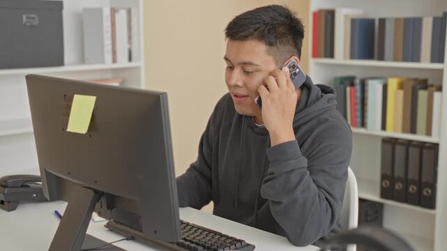 Man holds smartphone to ear while typing on computer at building desk, smiling and leaning forward with right hand on keyboard and sticky note on monitor; work focus.