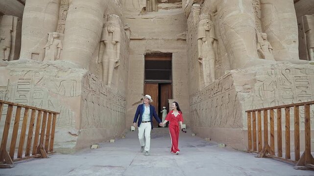 american young man traveler wearing white hat with his wife with red dress exploring ancient Egyptian abu simble temple with background of ramsses ii statues. Egypt.