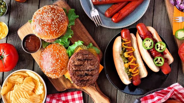 Summer BBQ food table scene with hot dog and hamburger buffet. Slow rotating zoom motion. Top view over a dark wood background.