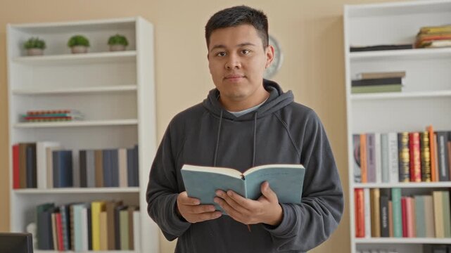 Man holding book in a library building, hands visible, wearing dark hoodie and standing by bookshelves while reading and gazing to the side; calm reflection.