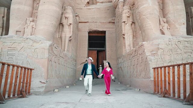 american young man traveler wearing white hat with his wife with red dress exploring ancient Egyptian abu simble temple with background of ramsses ii statues. Egypt.
