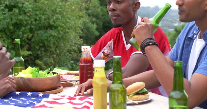 Vertical video: African American men at picnic table watching blue shirt lifting bottle, toasting
