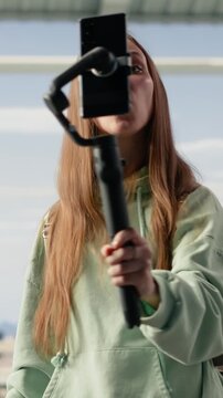 Vertical Video Girl content creator using phone on tripod to record vlog in airport lounge, capturing travel journey before boarding. Busy terminal illustrates social media presence and mobile filming