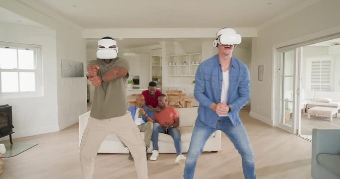 Diverse male friends playing VR for fun, two players wearing white headsets swinging arms at home