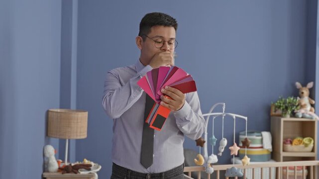 Man flipping a fan of pink and red color swatches with his hands beside a baby crib and mobile in a studio nursery; uncertainty choice.