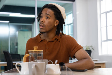 Fototapeta premium African American man 20s wearing knit hat rust polo sitting in office pouring from glass teapot