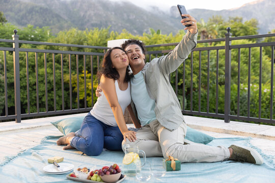 Romantic couple sitting on blue picnic blanket on rooftop terrace, taking selfie with smartphone