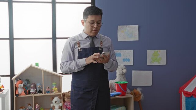 Man kindergarten teacher in apron tapping smartphone and pointing at screen in school building, surrounded by toys and drawings; calm guidance.