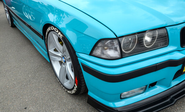 Bright turquoise BMW E36 features a custom front lip and "angel eye" headlights. Parked at a show, its silver multi-spoke wheels highlight a classic 90s tuning aesthetic