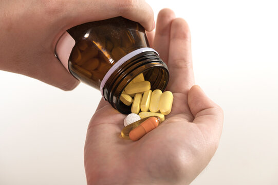 Close-up of hand holding various pills and capsules pouring from brown medicine bottle. Concept of healthcare, medication, treatment, vitamins, and pharmaceutical.