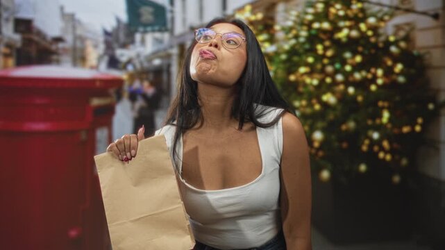 Woman holding paper bag and sticking tongue out on street with christmas tree and red postbox; playful defiance.