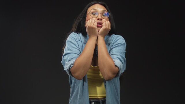 Woman clutching hands to cheeks and biting her lip wearing denim shirt and yellow tanktop in studio; anxiety tension anticipation.