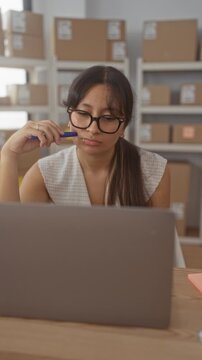 Woman latina at laptop holds pen to lip in building parcel office with shelves of packages; small business planning pensive.