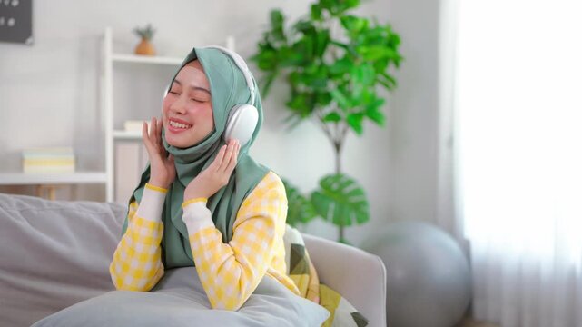 Asian young Muslim woman listen to music while sitting on sofa in house. 
