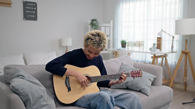 Asian young man playing an acoustic guitar in cozy living room at home. 