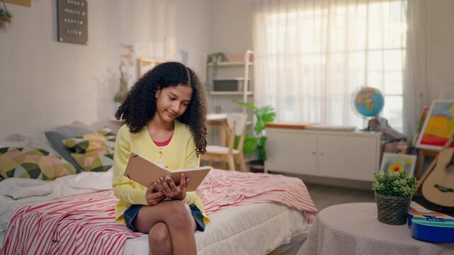 African American teenage girl sitting on comfort bed and reading a book.
