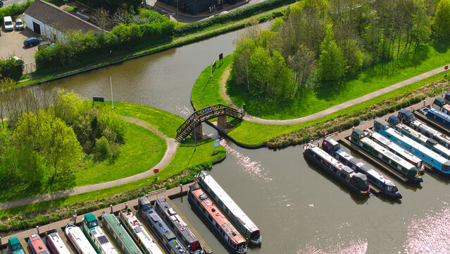 Aerial view of Barton Marina featuring narrowboats moored in rows and a wooden footbridge over a canal junction surrounded by lush green trees in Barton-under-Needwood, England, United Kingdom.