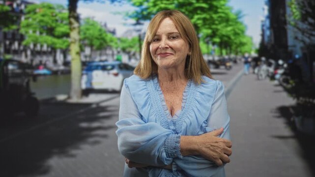 Middle age hispanic woman with bare hands crossing arms on a tree lined street by a canal and parked cars; maturity confidence.