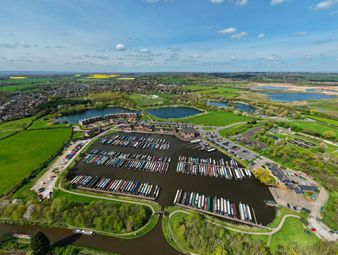 Aerial view of Barton Marina with narrowboats docked in the basin, surrounded by lush green fields and lakes under a blue sky in Barton-under-Needwood, England, United Kingdom.