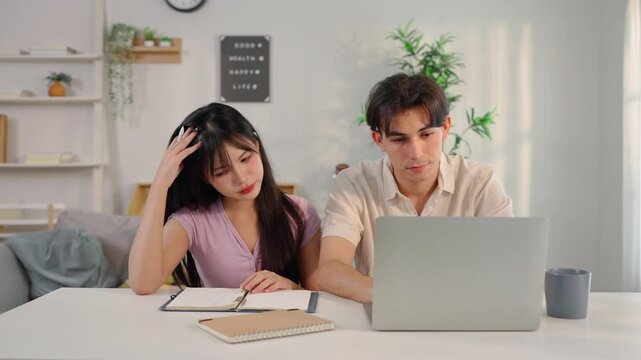 A serious young couple sits at a table and working on a laptop in house. 