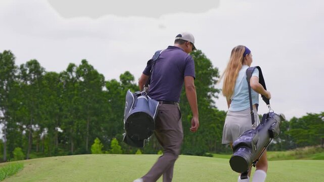 Asian young couple golfer walk across lush green fairway with golf club.