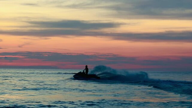 Athletic man silhouette riding a fast jet ski at sunset on Phuket beach. Extreme water sports action against a vibrant orange sky, tropical summer adventure and freedom concept.