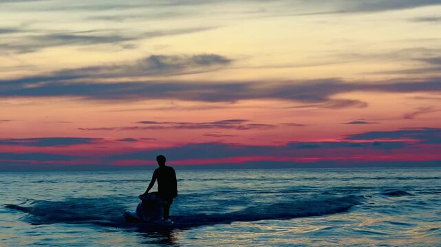 Athletic man silhouette riding a fast jet ski at sunset on Phuket beach. Extreme water sports action against a vibrant orange sky, tropical summer adventure and freedom concept.