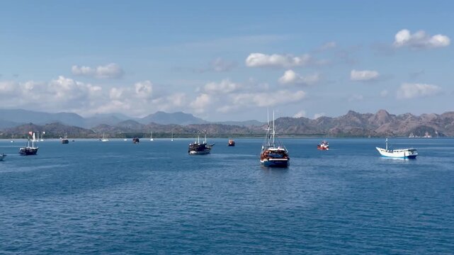 Wide tracking shot of various traditional indonesian wooden boats and yachts floating on calm blue ocean in labuan bajo