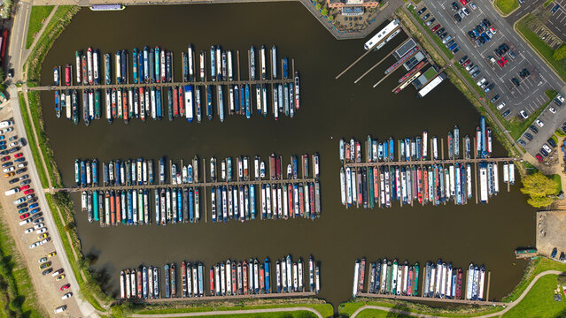 Aerial view of Barton Marina with rows of colorful narrowboats moored at wooden jetties, surrounding parking lots and green grassy banks Barton-under-Needwood, England, United Kingdom.