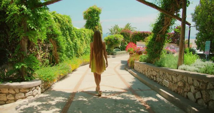 Young woman walking down a sunny garden pathway surrounded by lush summer plants in Capri