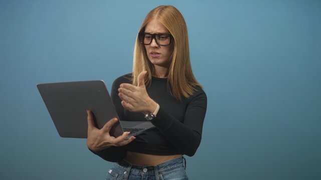Woman holding laptop with left hand and gesturing with right hand while looking puzzled, wearing glasses and crop top in studio; frustration.
