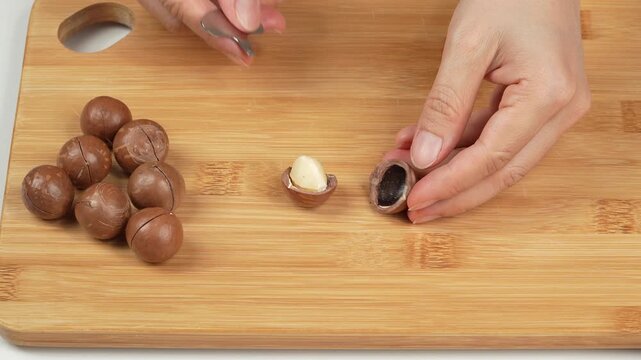 Hands opening macadamia nut with metal opener tool on wooden board.