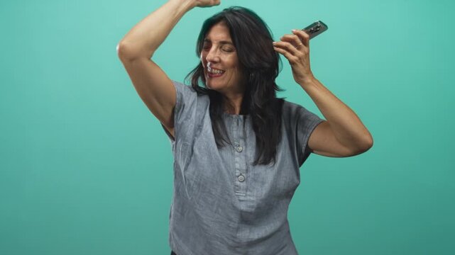 Woman holds smartphone and raises arm while smiling with eyes closed in studio; celebration joy music.