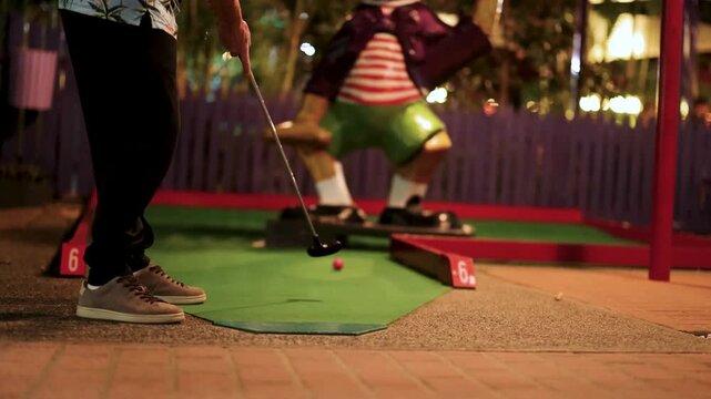 Person playing miniature golf at night, putting ball on green course with obstacle.