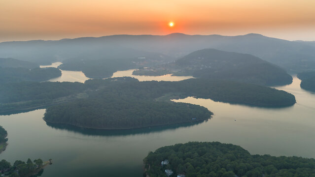Aerial view of Tuyen Lam Lake in Da Lat, Vietnam at sunrise, where the golden sun rises above layers of misty mountains. The winding lake reflects soft morning light, creating a tranquil and dreamy at