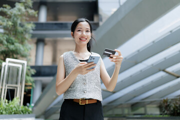 A woman smiles outdoors while holding a smartphone and a credit card, appearing to make an online transaction in a modern urban setting. © Minttita