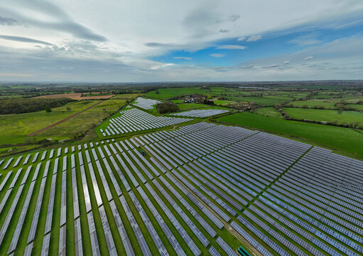 Aerial view of Packington Solar Farm featuring rows of photovoltaic panels set amidst lush green fields and rural landscapes under a dramatic cloudy sky in Packington, England, United Kingdom.