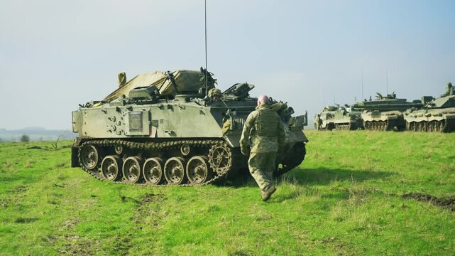 British Army soldier walking next to an armoured tracked vehicle on open grassland during a military training exercise on Salisbury Plain, United Kingdom