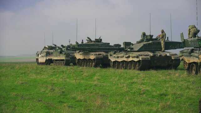 Multiple armoured military tanks with soldiers positioned on top during a training exercise on open grassland terrain in Salisbury Plain, United Kingdom